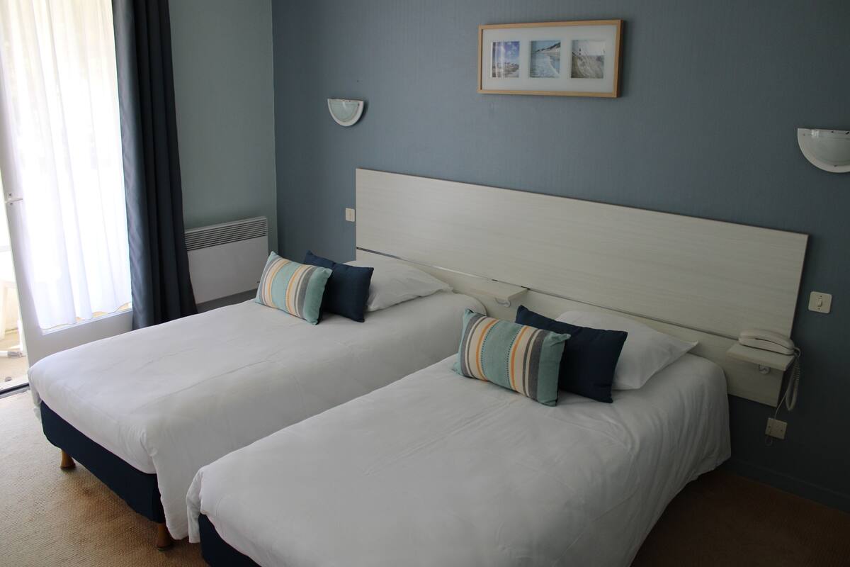 A hotel room with two beds, a bedside table with a lamp, a framed picture on the wall, and a window with curtains.