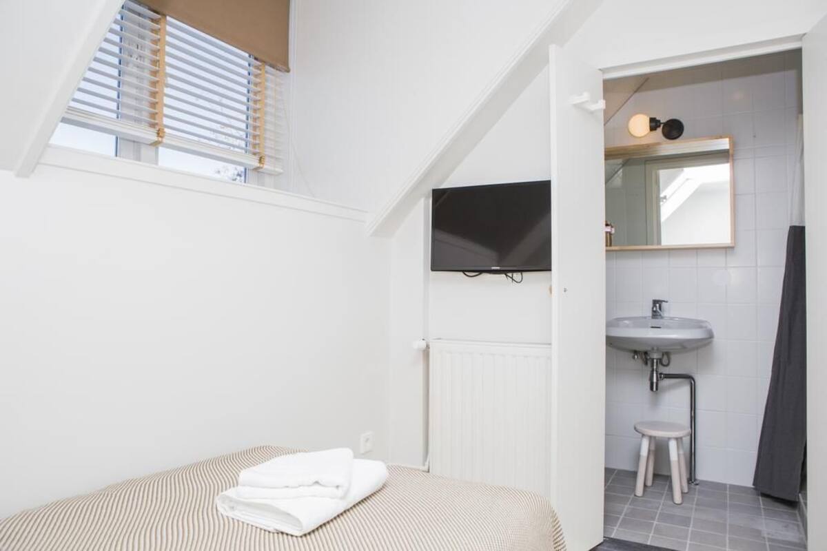 Un dormitorio con cama, ventana con persianas, televisor montado en la pared, baño con lavabo y espejo, y un radiador.