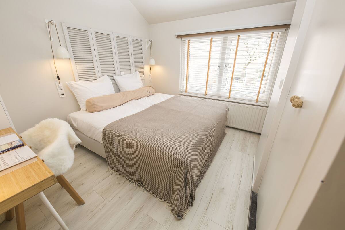 Un dormitorio con cama, mesita de noche, lámpara y ventana con persianas.