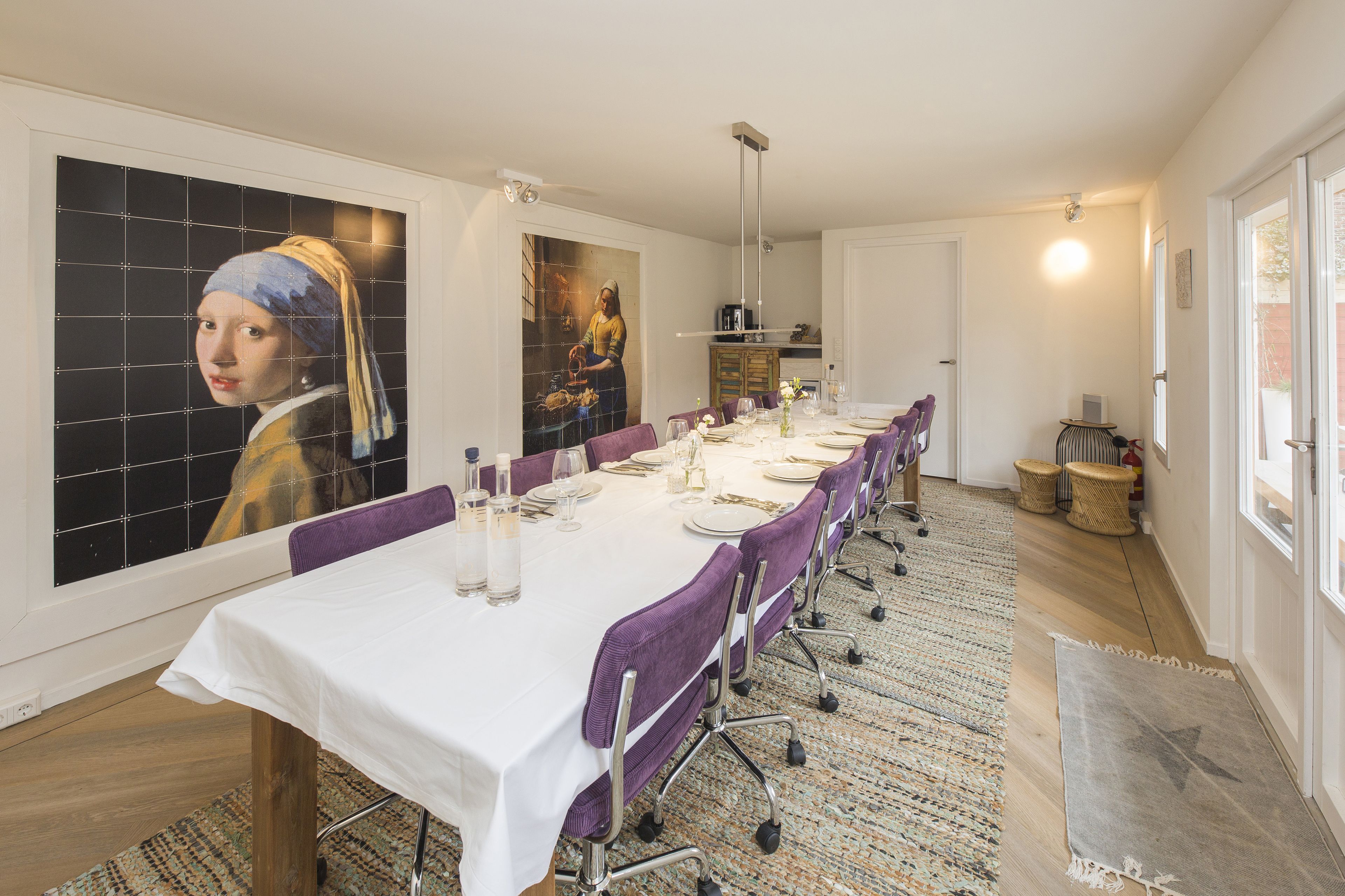 A dining area with a long table set for six, purple chairs, a white tablecloth, and a large painting on the wall.