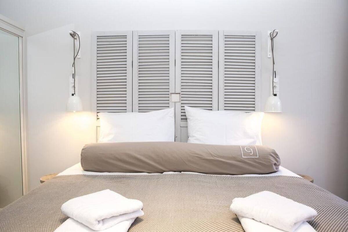 Una cama bien hecha con ropa de cama blanca y beige, dos lámparas de noche y una ventana con persianas.