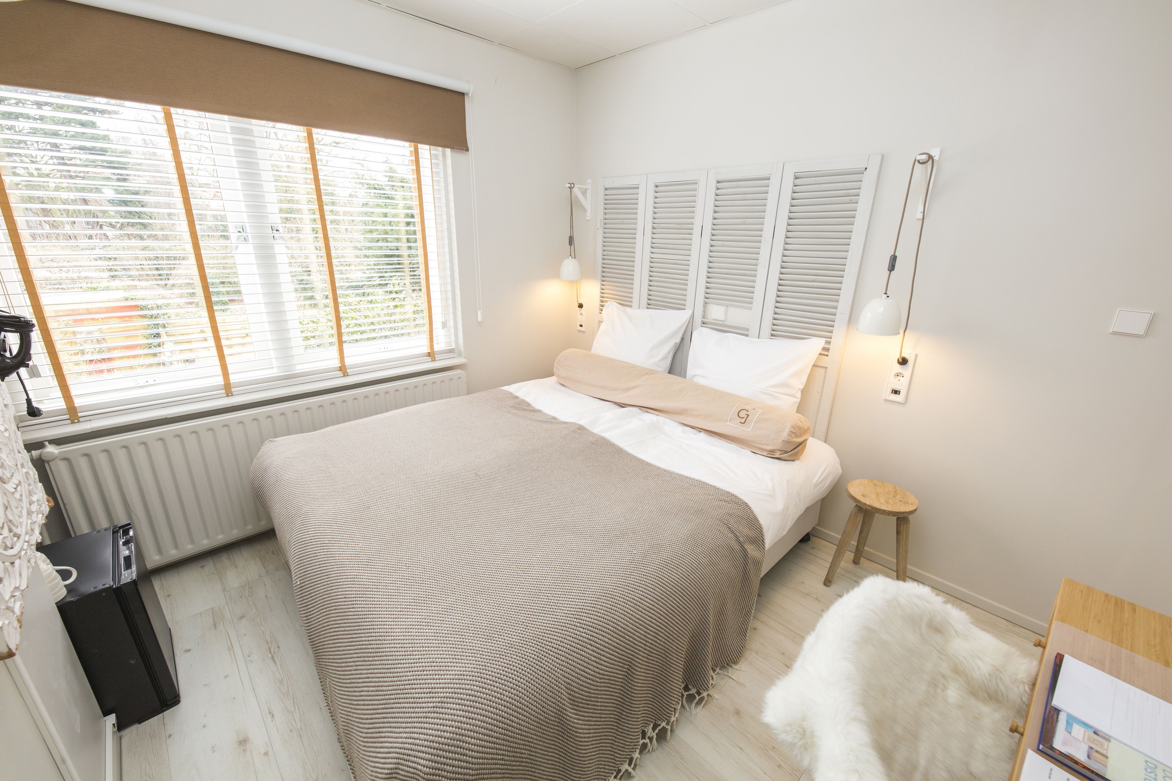 A bedroom with a large bed, a radiator, a window with blinds, a bedside table, and a lamp.