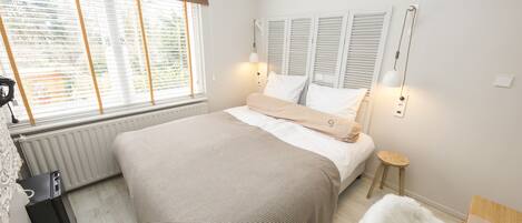 A bedroom with a large bed, a radiator, a window with blinds, a bedside table, and a lamp.
