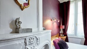 Un dormitorio con una chimenea blanca, una cama con almohadas moradas y grises, una mesita de noche con una lámpara y una ventana con cortinas.