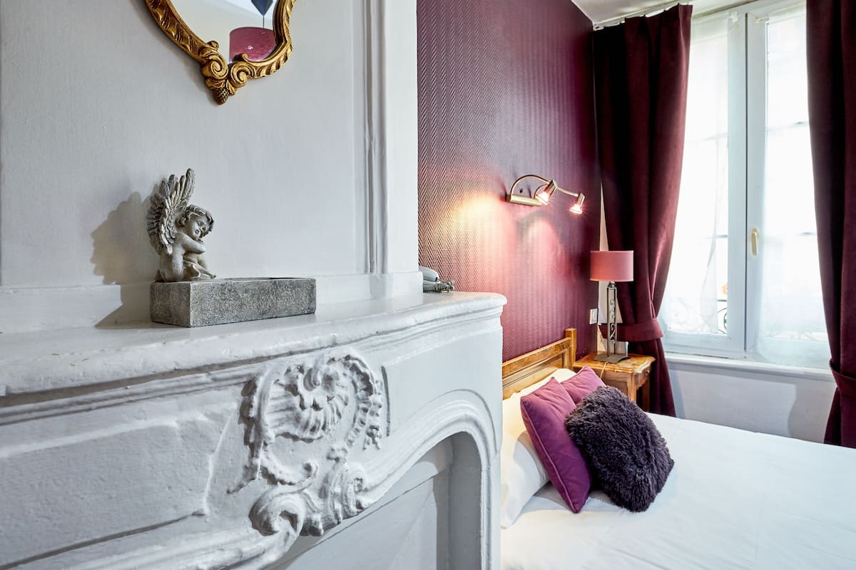 A bedroom with a white fireplace, a bed with purple and gray pillows, a nightstand with a lamp, and a window with curtains.