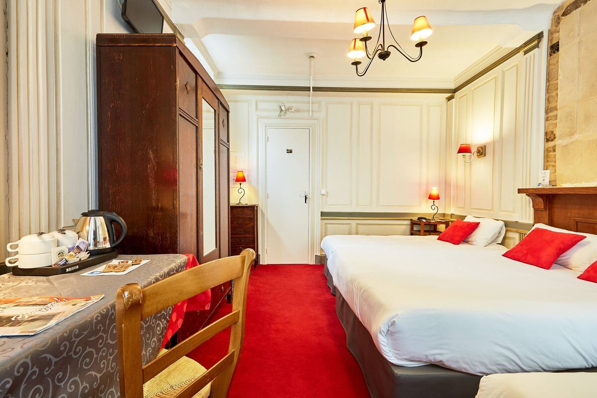 A hotel room with a wooden wardrobe, a table with a tray of tea, a bed with red pillows, and a chandelier.