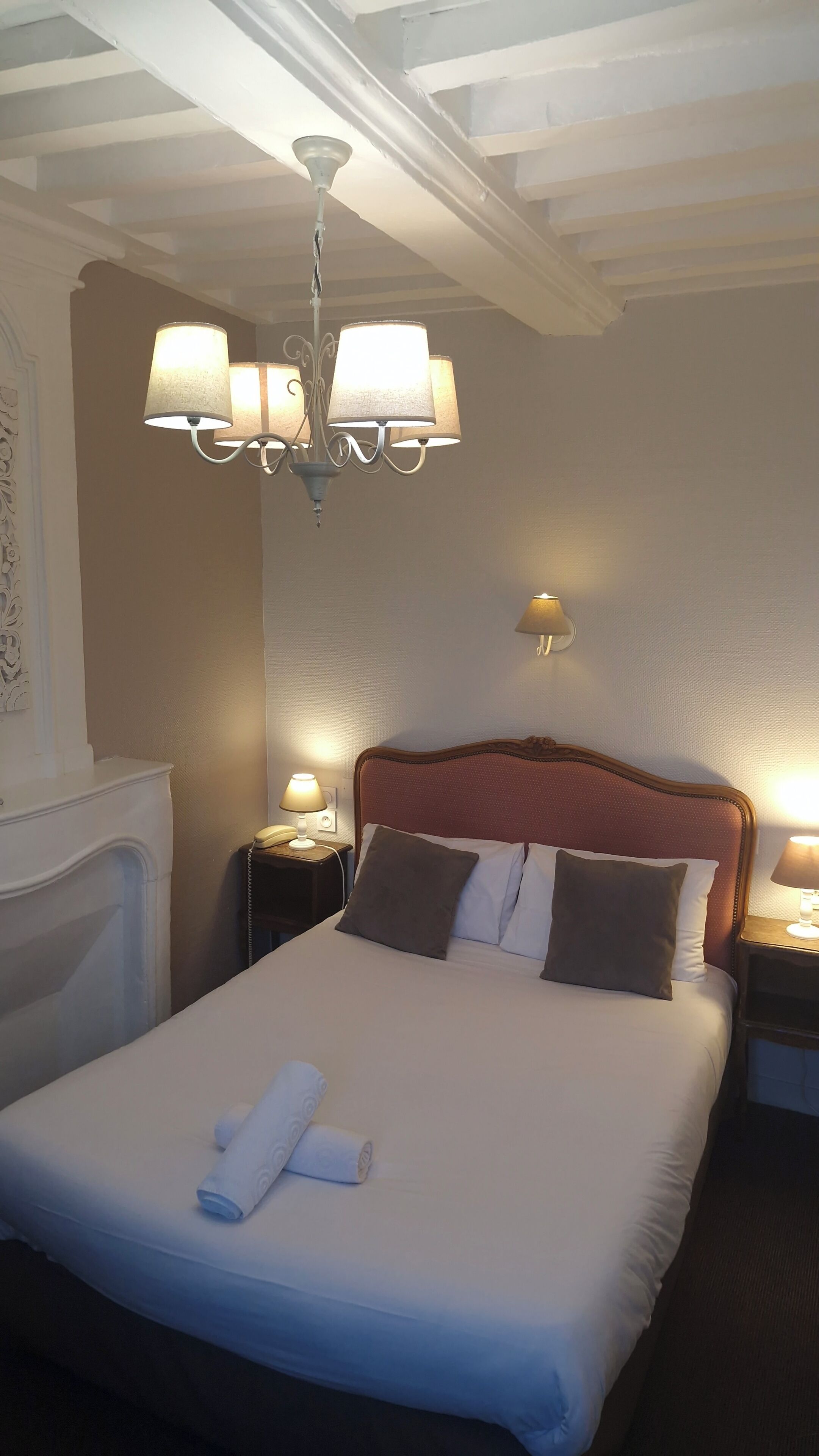 A hotel room with a large bed, a chandelier, a bedside table with a lamp, and a wall-mounted light.