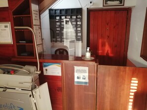 Ett välkomstområde med ett skrivbord i trä, en stol, en dispenser för handsprit, en anslagstavla med information och en dörr med en skylt som uppmanar besökare att använda desinfektionsmedel vid entrén.