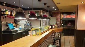 A restaurant interior with a long wooden counter, black bar stools, a cash register, and a brick wall with a fireplace. - Hostal Jaime I (Huesca)