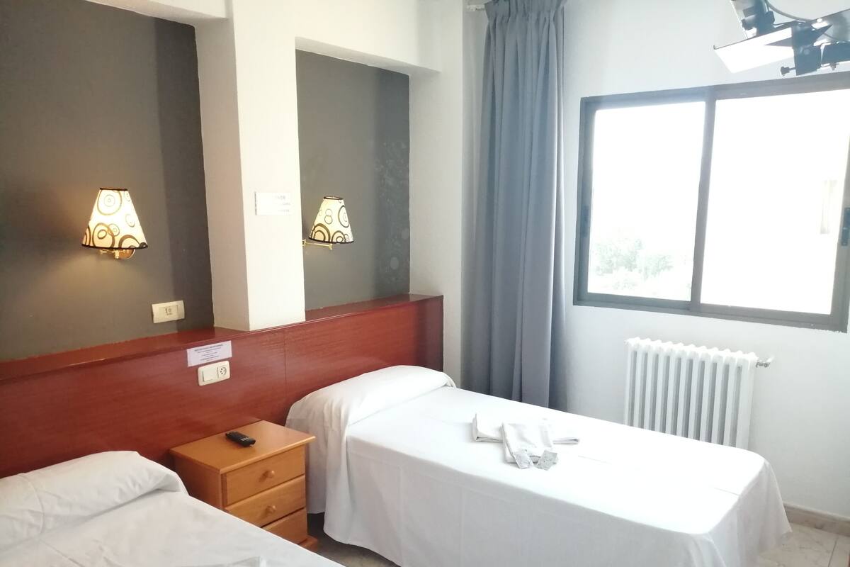 A hotel room with two single beds, a wooden nightstand, a mirror, a window with curtains, and wall-mounted lamps.
