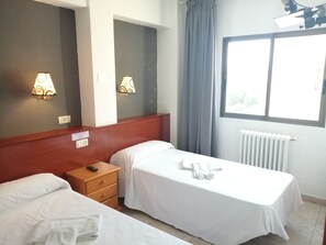 Ett hotellrum med två separata sängar, ett nattduksbord i trä, en spegel, ett fönster med gardiner och väggmonterade lampor.
