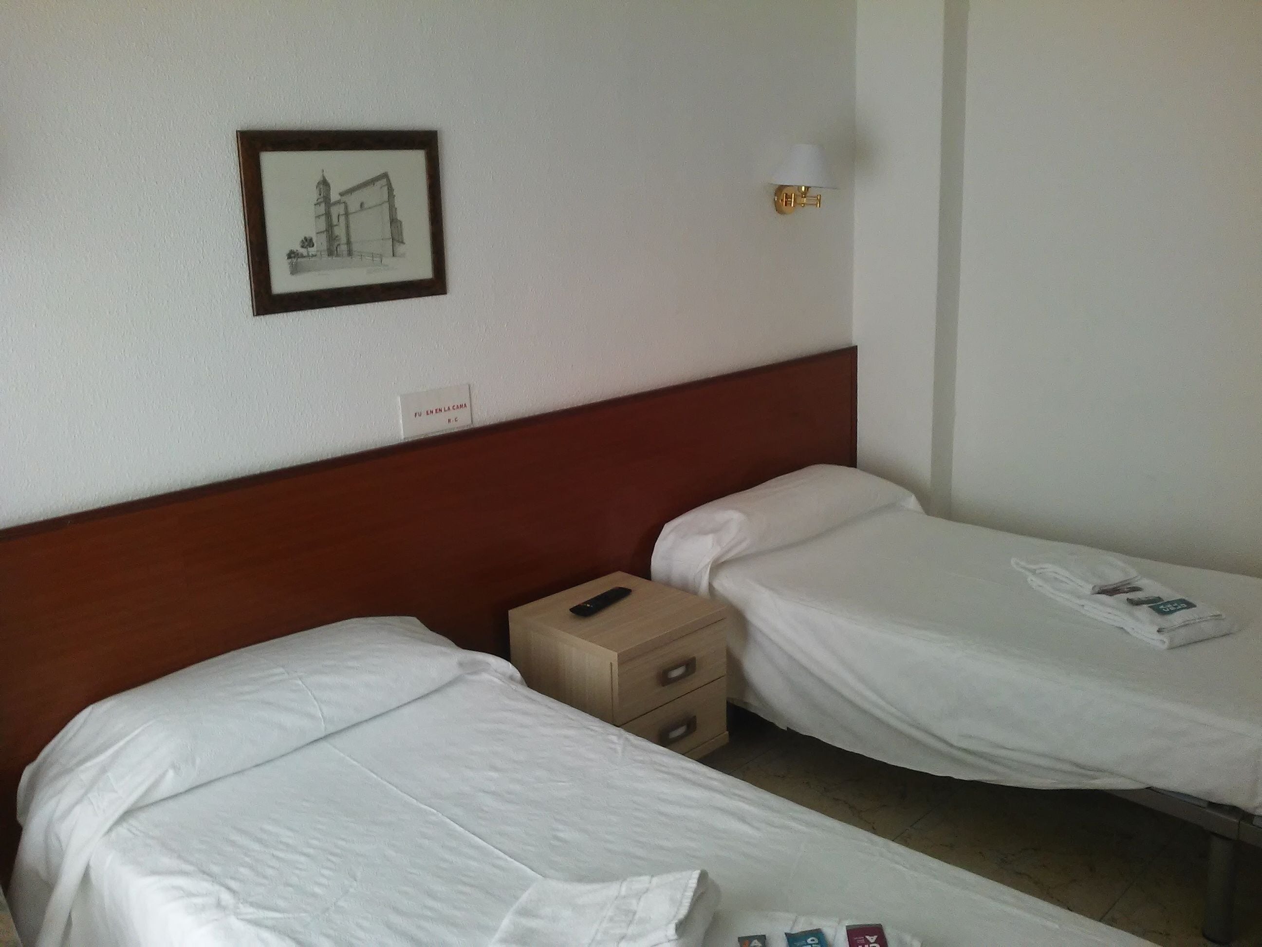 Habitación de hotel con dos camas individuales, cabecera de madera, mesita de noche con lámpara, cuadro en la pared y un cartelito sobre la puerta.