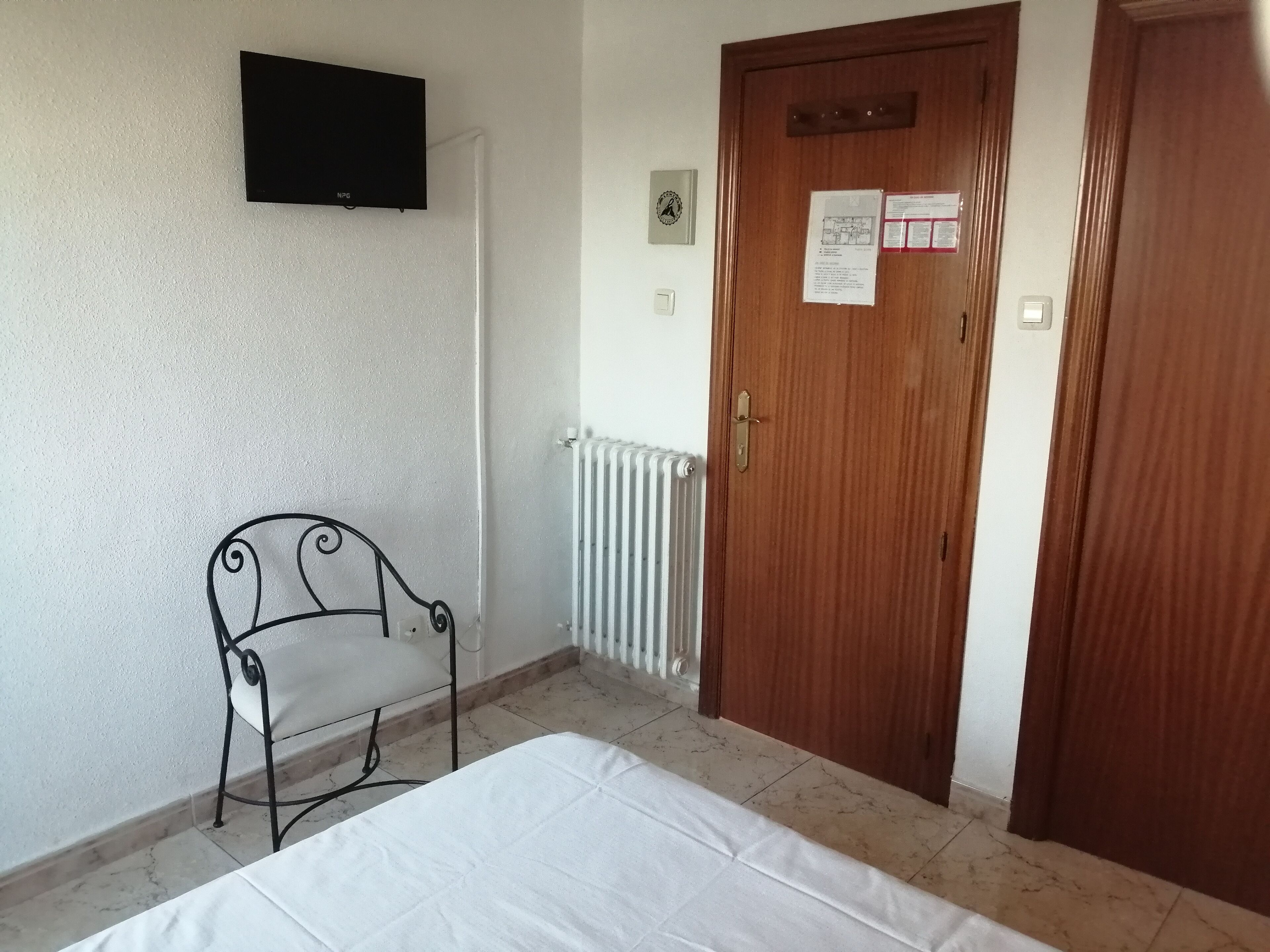 A room with a bed, a chair, a wooden door, a radiator, and a TV mounted on the wall.