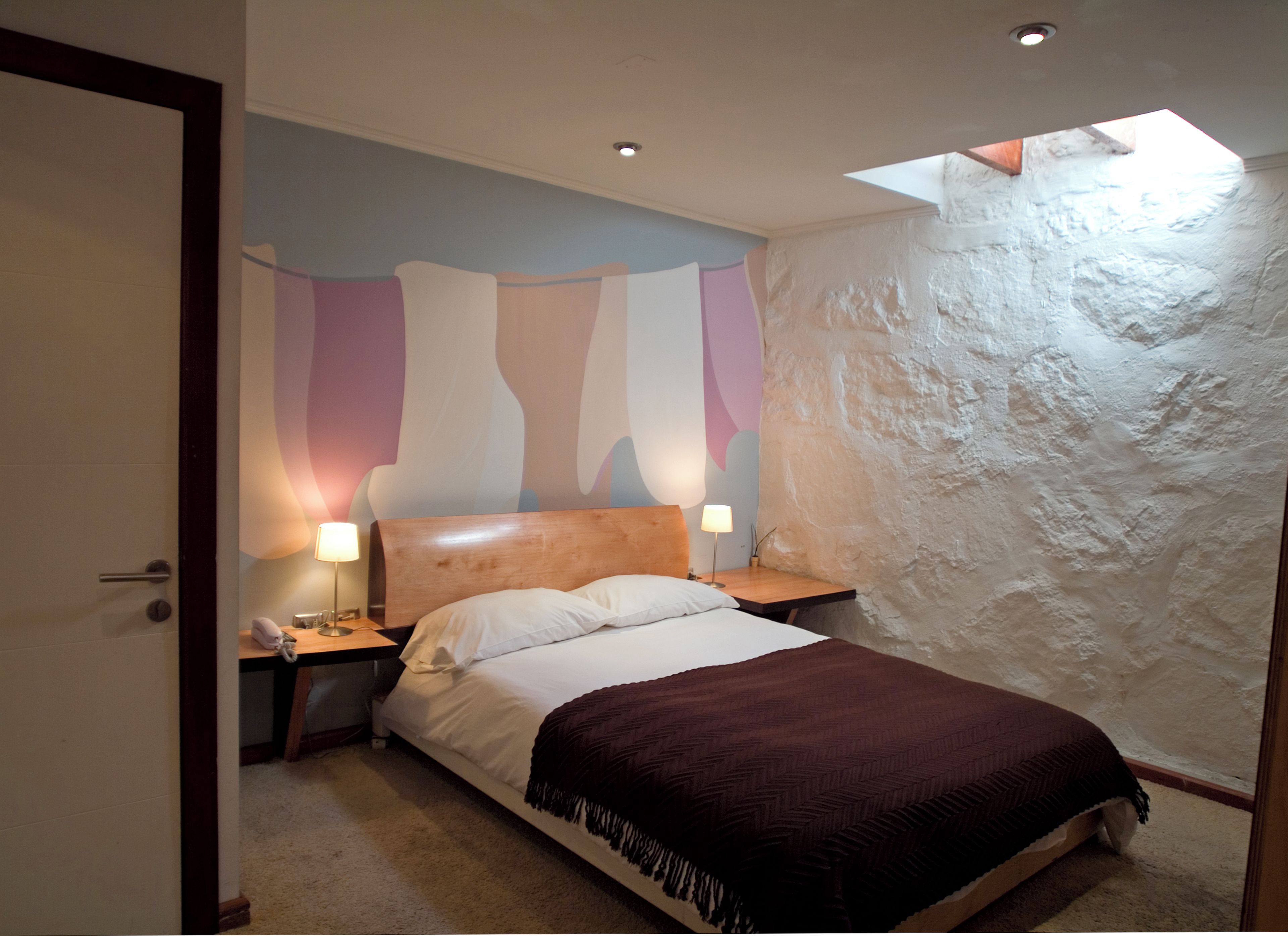 Un dormitorio con una cama, dos mesitas de noche, una lámpara de noche, una pared con diseño texturizado y un tragaluz.