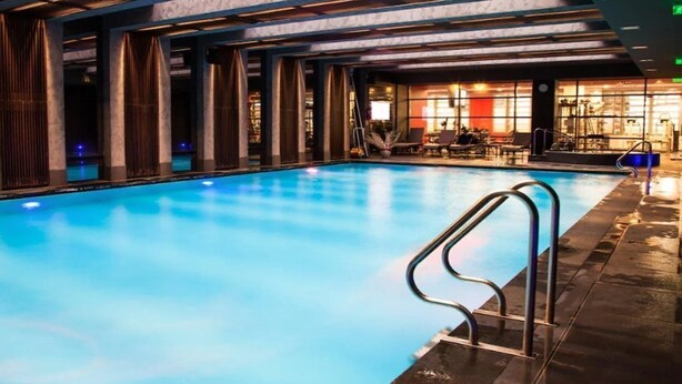 A swimming pool with a clear blue water surface, surrounded by a modern interior with a patterned wall and a seating area with chairs and tables. - City Gardens Apartments (Budapest)
