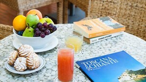 A table with a fruit bowl, pastries, drinks, and books in a cozy indoor setting with wicker furniture and potted plants. - Hotel Sabrina (Castiglione della Pescaia)