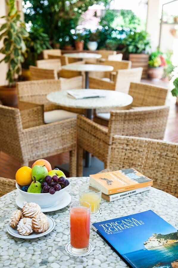 A table with a fruit bowl, pastries, drinks, and books in a cozy indoor setting with wicker furniture and potted plants. - Hotel Sabrina (Castiglione della Pescaia)