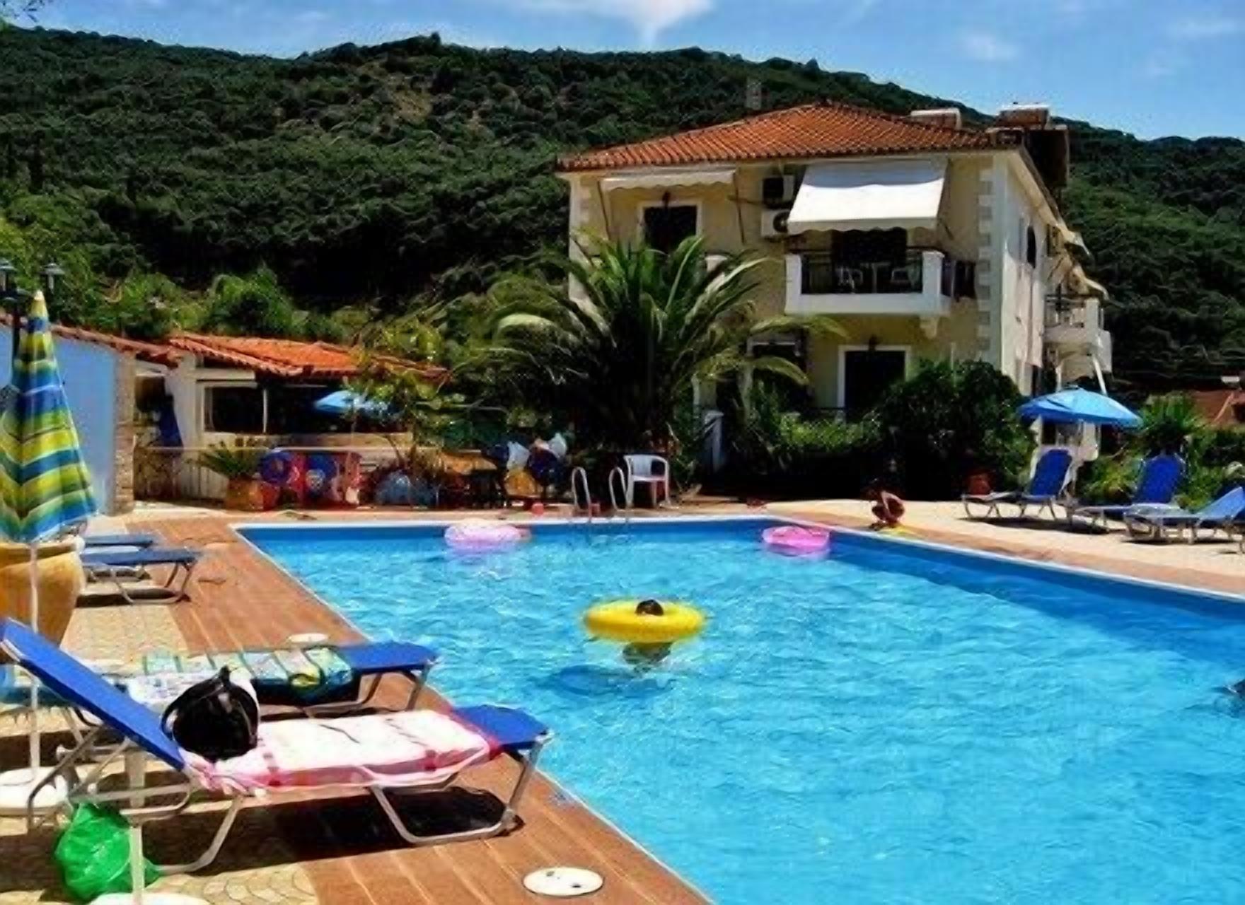 A swimming pool with lounge chairs and umbrellas, surrounded by greenery and a building.