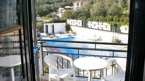 A balcony with a pool, white plastic chairs, and a view of a mountainous area with buildings. - Harmony Resort (Parga)