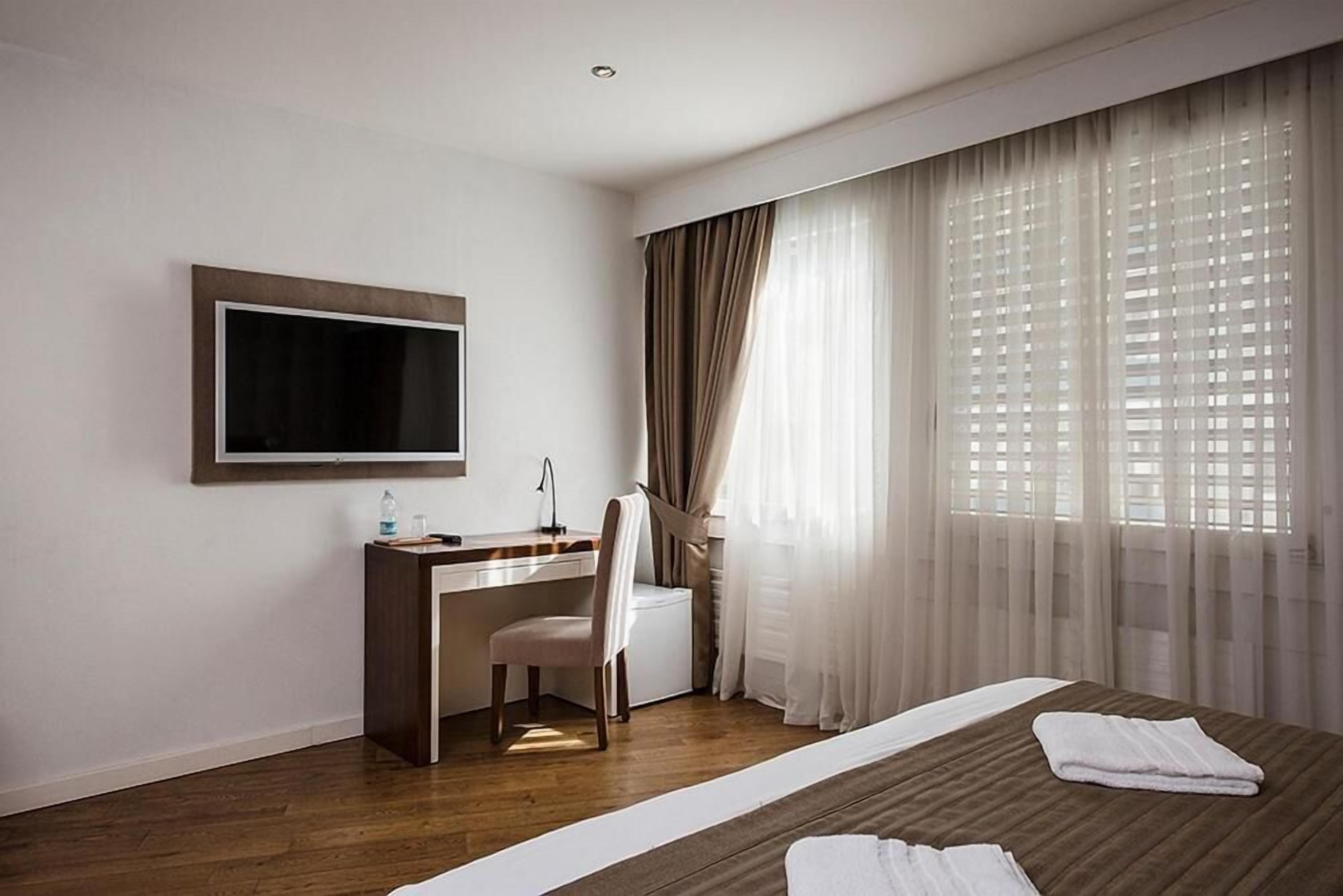 Ein Hotelzimmer mit Bett, Schreibtisch, Sessel, Fernseher und großen Fenstern mit Vorhängen.