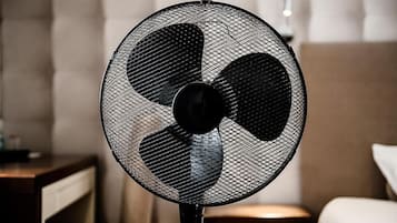 Ein Standventilator mit Schutzgitter und drei Flügeln, platziert in einem Zimmer mit Bett und Nachttisch.