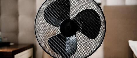 A standing fan with a mesh grill and three blades, positioned in a room with a bed and a nightstand.