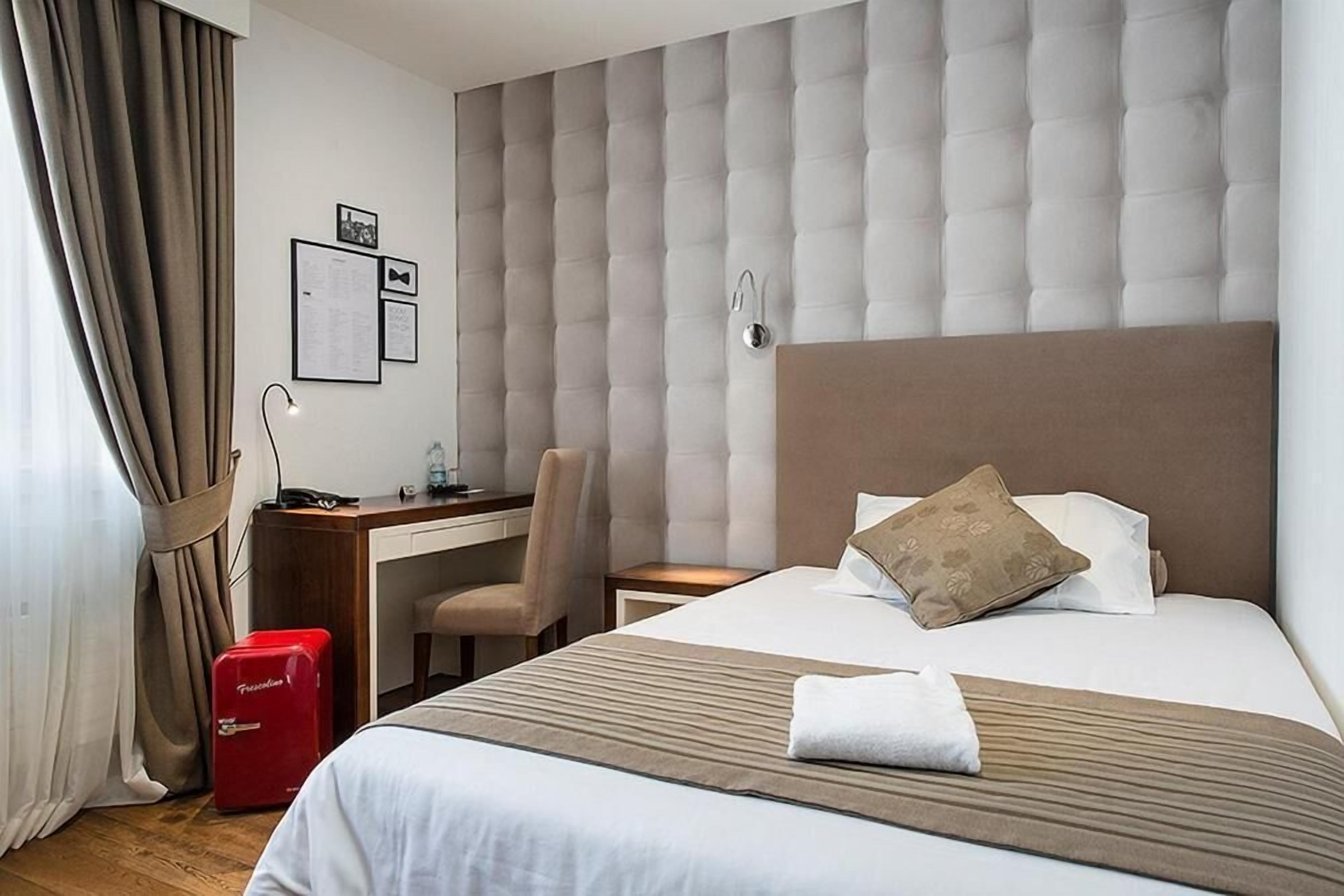 A hotel room with a bed, a desk, a chair, and a red suitcase.