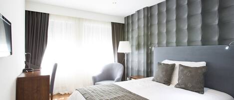 A modern hotel room with a large bed, a desk, a chair, and a window with curtains.