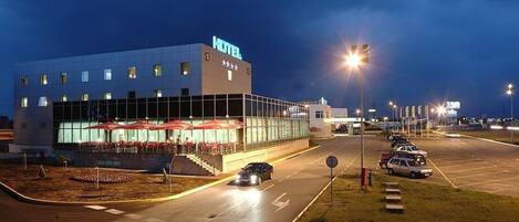A modern hotel with a glass facade, outdoor seating area, and a parking lot.