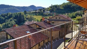 On voit des bâtiments en pierre à étages, surmontés de toits en tuiles rouges, un balcon avec une balustrade en métal, et un paysage montagneux en arrière-plan.