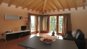 Un salón amplio con techo de madera, un sofá negro, una mesa de comedor con un bol de fruta y un televisor sobre un soporte blanco.