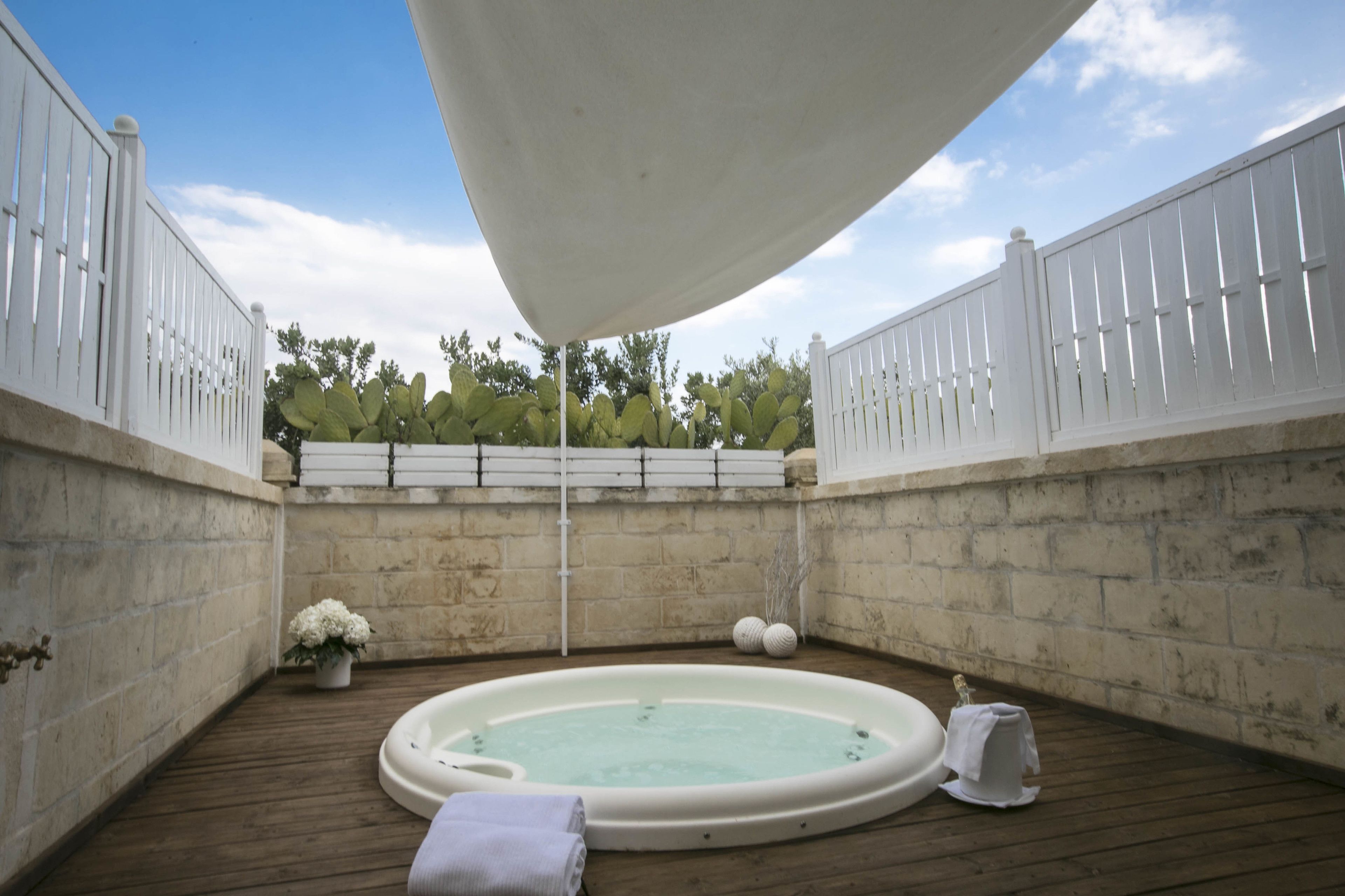 A hot tub in a covered outdoor area with a wooden deck, white fencing, and a brick wall.