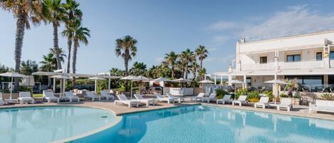 Un espace piscine agrémenté de chaises longues et de parasols, entouré de palmiers et bordé d’un bâtiment doté de balcons.