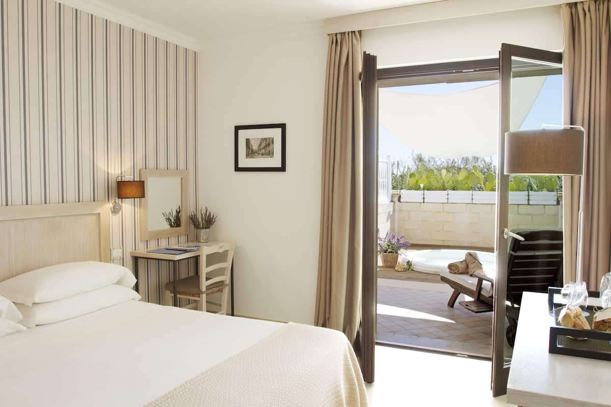 Une chambre d’hôtel avec un lit, un bureau doté d’une lampe, une chaise, un tableau au mur et une porte ouverte donnant sur un espace extérieur avec une table et des chaises.