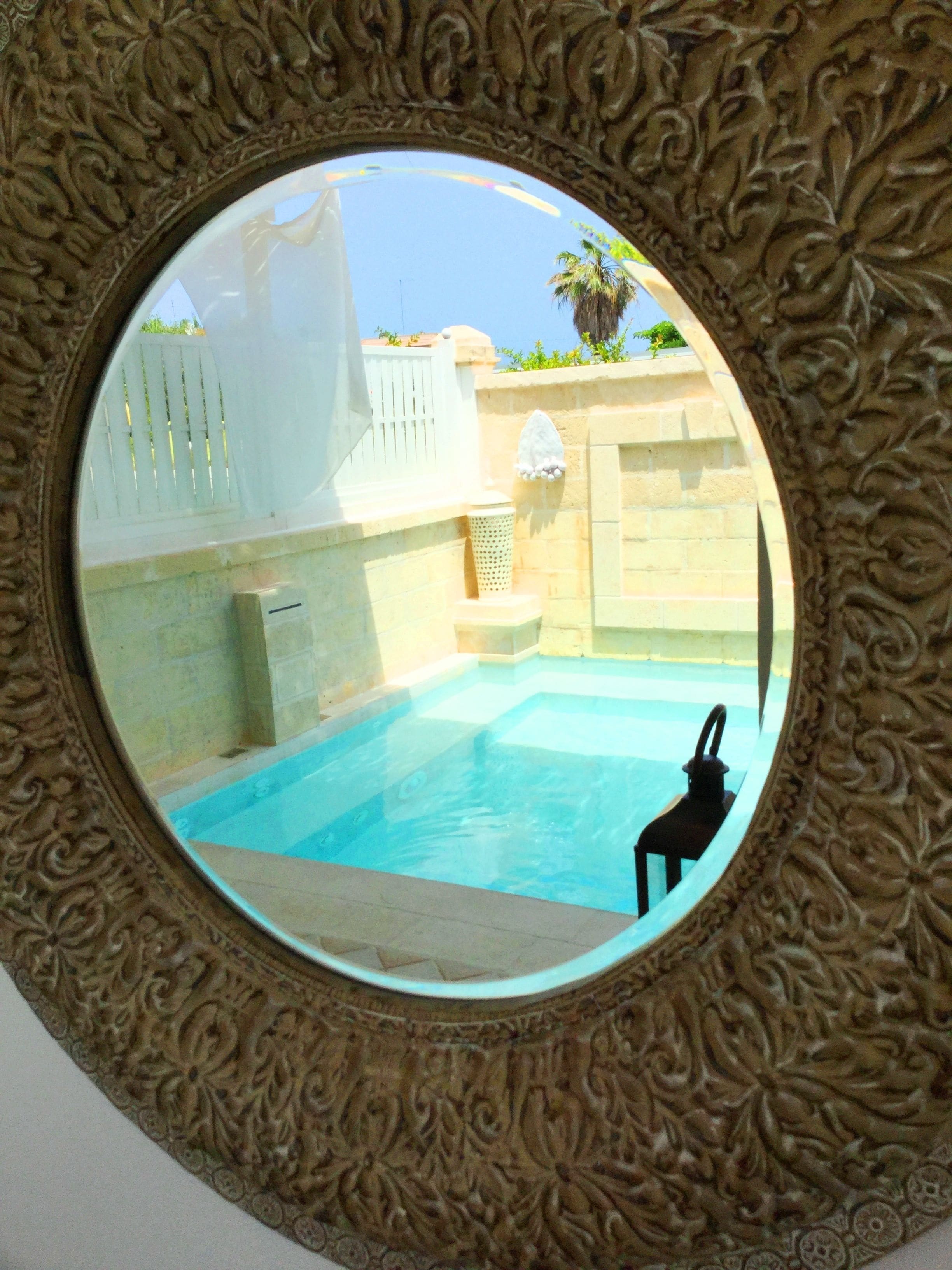 A circular mirror with ornate carvings, reflecting a swimming pool area with a tiled floor, a white fence, and a palm tree.