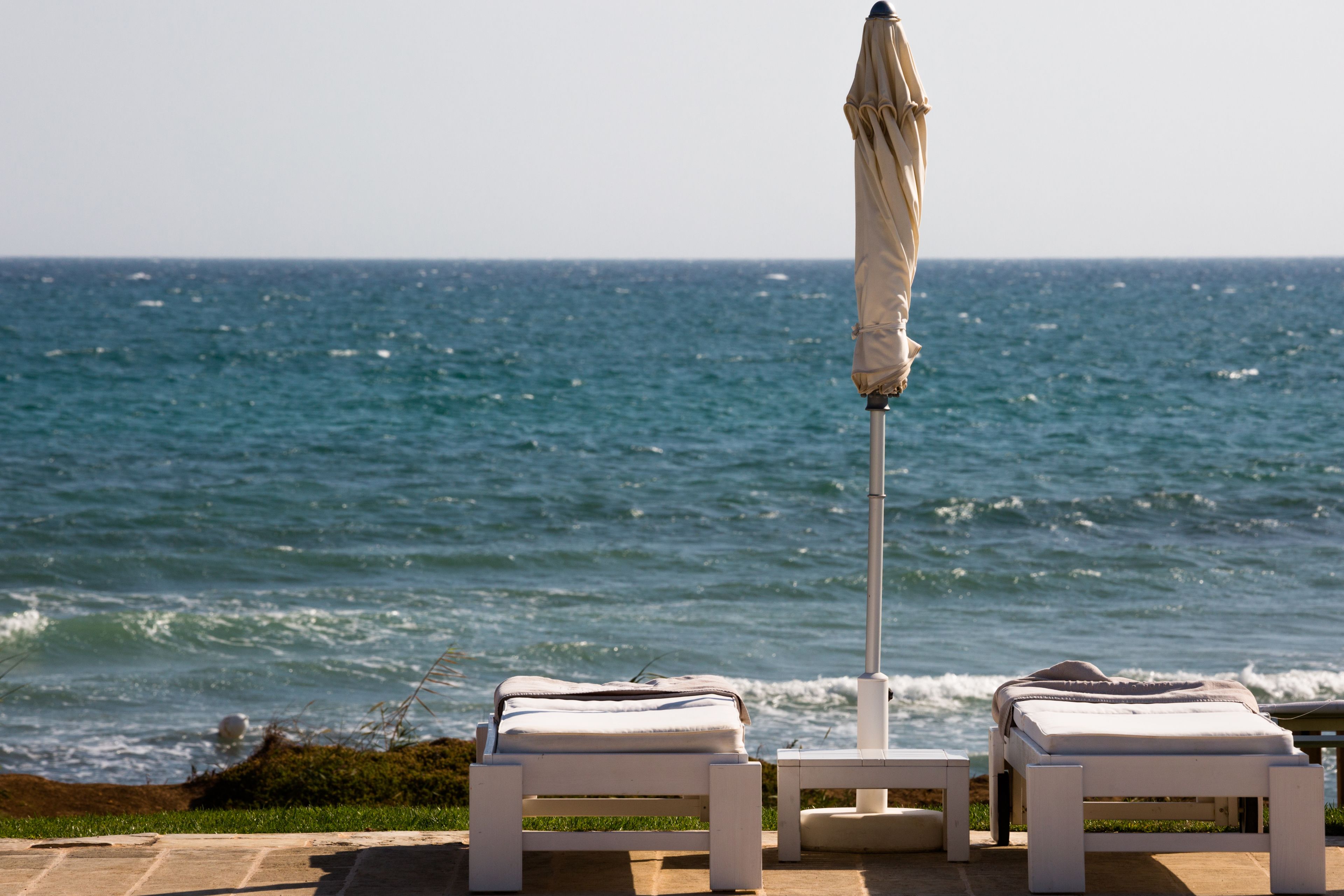 Two white lounge chairs with a beige umbrella on a patio overlooking the ocean.