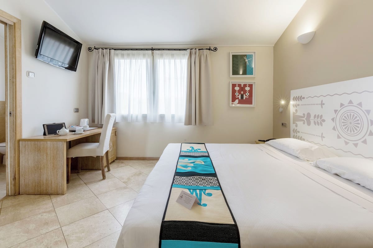 A hotel room with a bed, a desk with a chair, a television mounted on the wall, and a window with curtains.