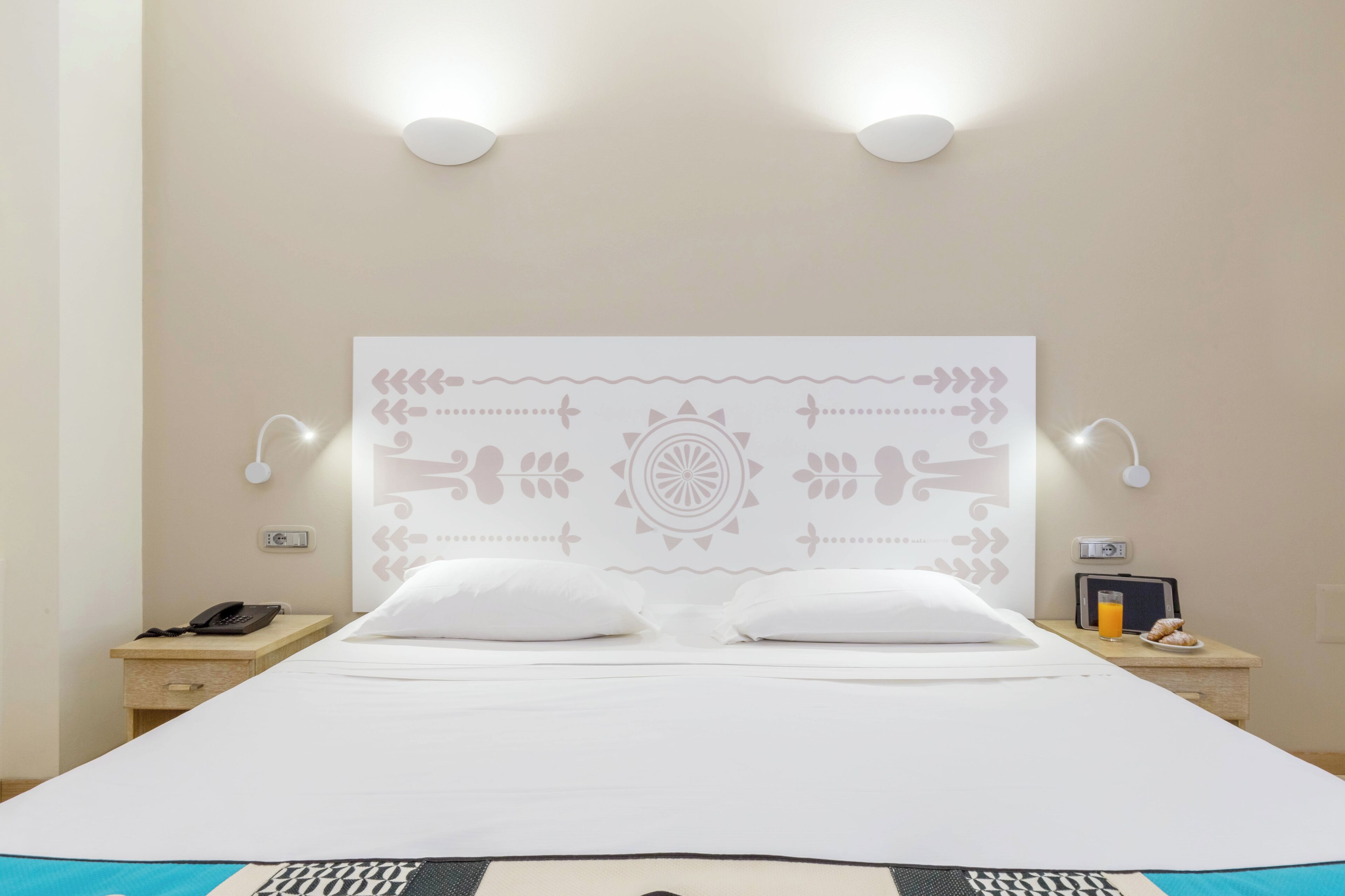 A neatly made bed with white linens, a decorative headboard with a central pattern, bedside lamps, a nightstand with a phone, and a small table with a glass of orange juice and pastries.