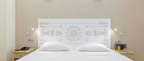 A neatly made bed with white linens, a decorative headboard with a central pattern, bedside lamps, a nightstand with a phone, and a small table with a glass of orange juice and pastries.