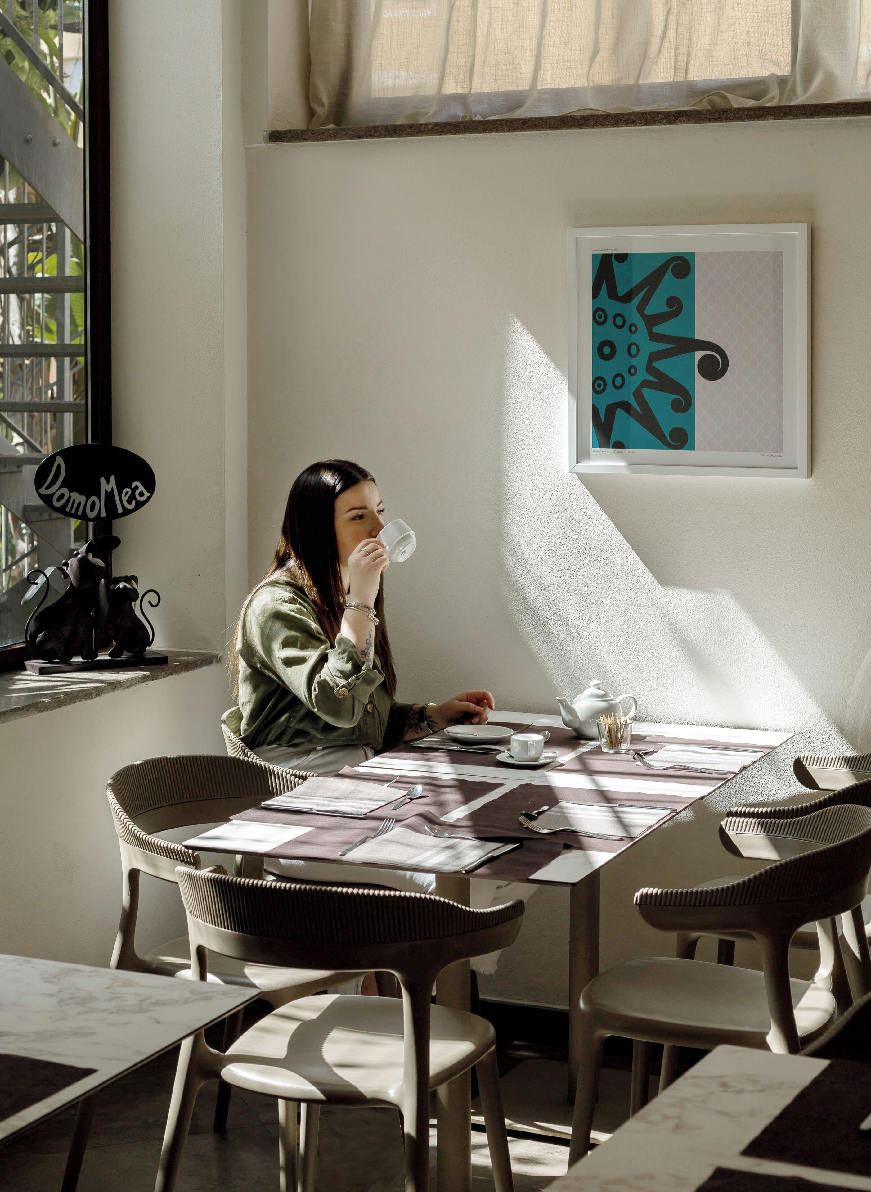 A person sitting at a table with a cup, in a room with a window, a framed artwork, and a sign on the wall.