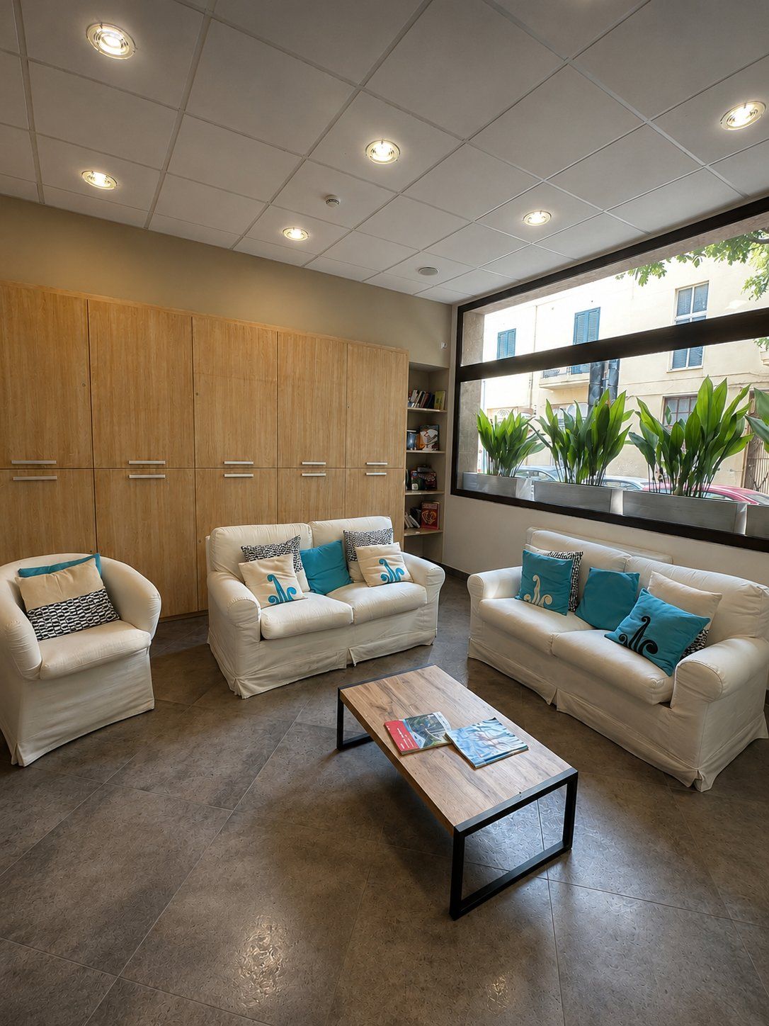 A modern living room with three white sofas, a wooden coffee table, and a large window with plants.