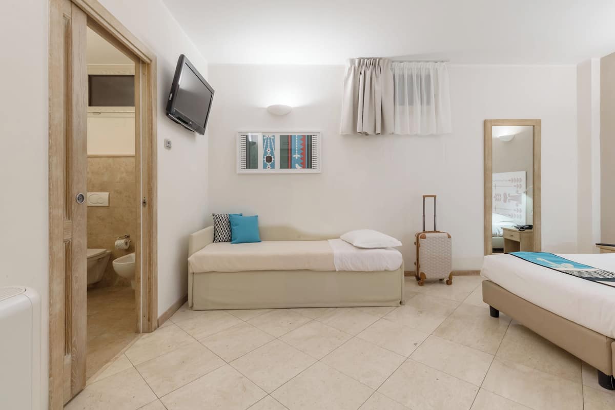 A hotel room with a bed, a sofa, a television, a mirror, and a suitcase.