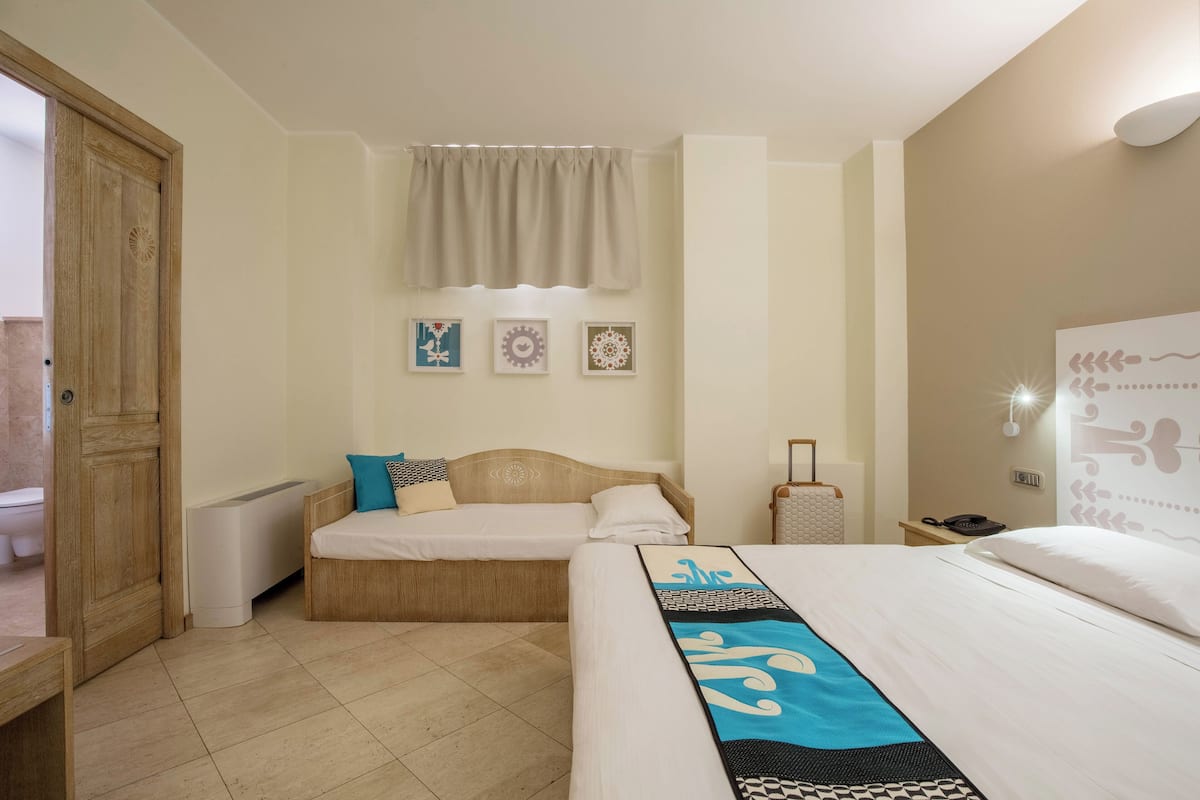 A hotel room with a large bed, a smaller bed, a wooden wardrobe, a radiator, a bedside table with a lamp, and a wall-mounted phone.