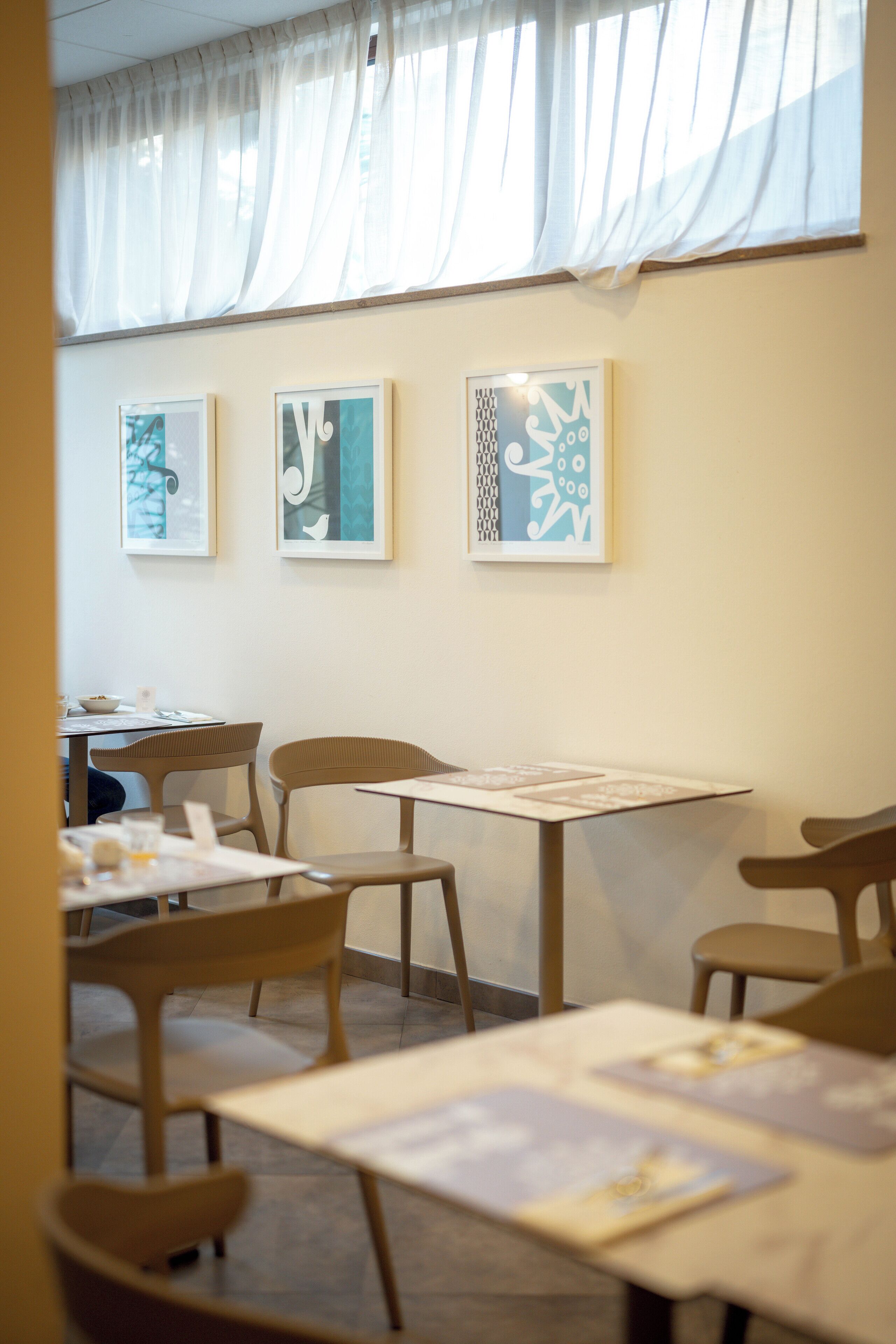 A dining area with tables and chairs, framed artwork on the wall, and a window with sheer curtains.
