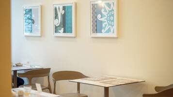 A dining area with tables and chairs, framed artwork on the wall, and a window with sheer curtains.