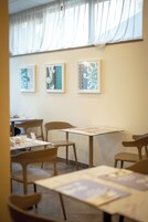 A dining area with tables and chairs, framed artwork on the wall, and a window with sheer curtains.