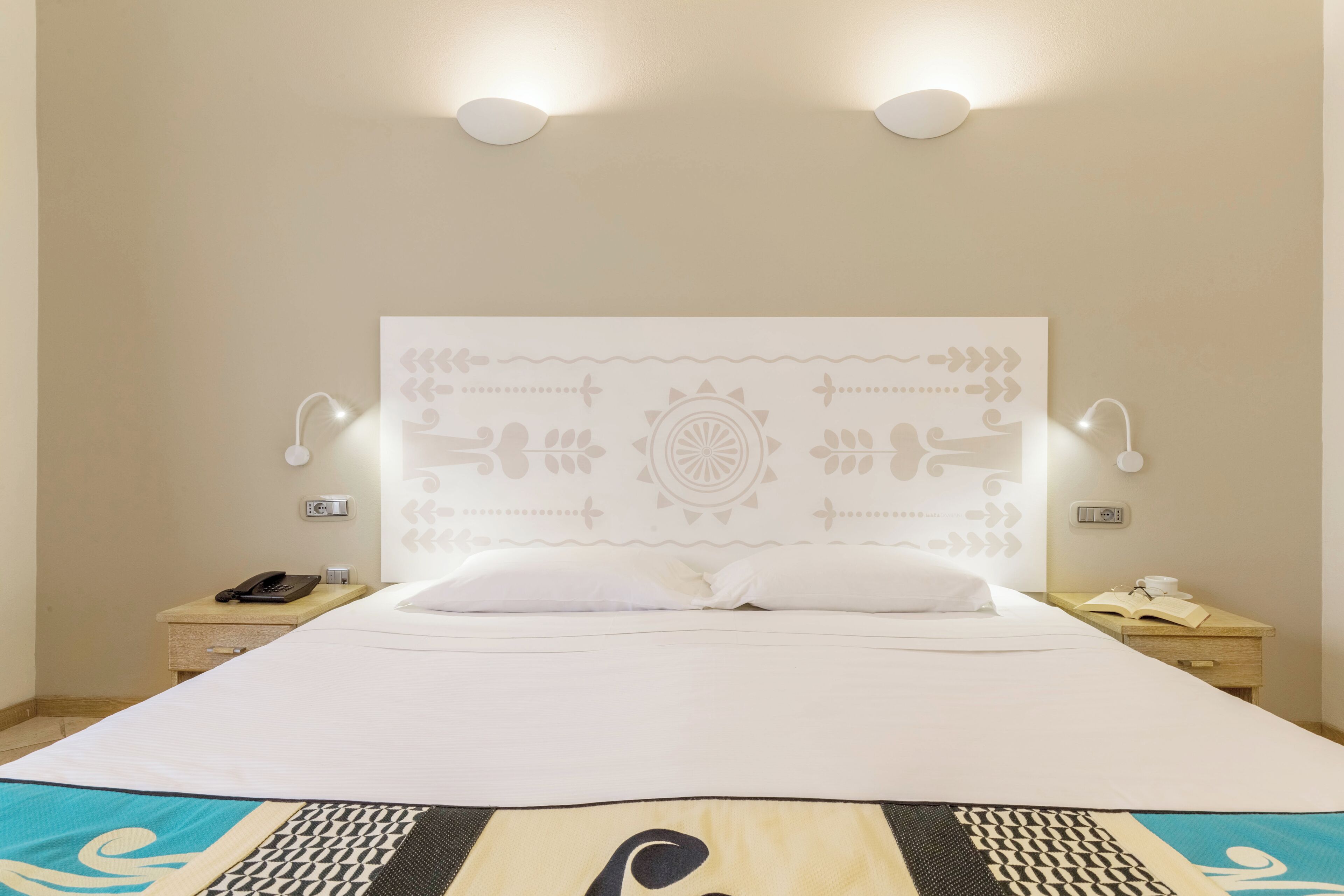 Una cama bien hecha con un cabecero estampado, lámparas de noche, un teléfono junto a la cama y una mesita con un libro y una taza.
