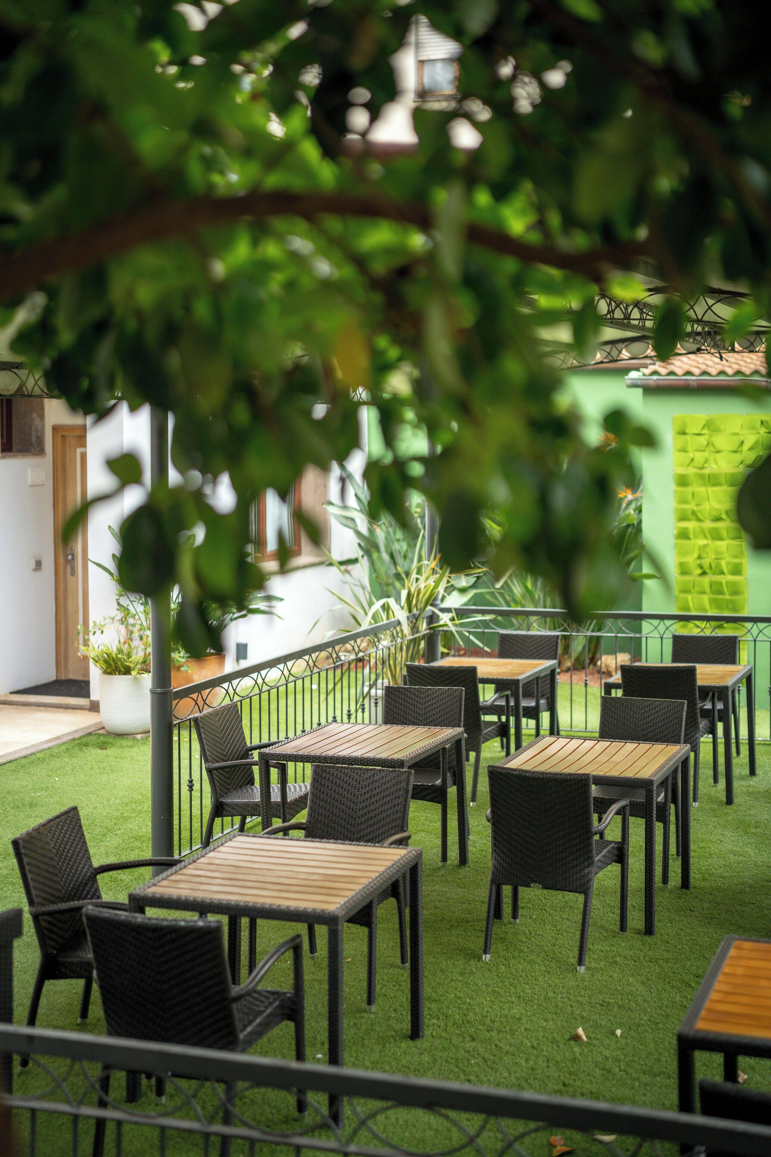 An outdoor seating area with tables and chairs, surrounded by greenery and a building in the background.