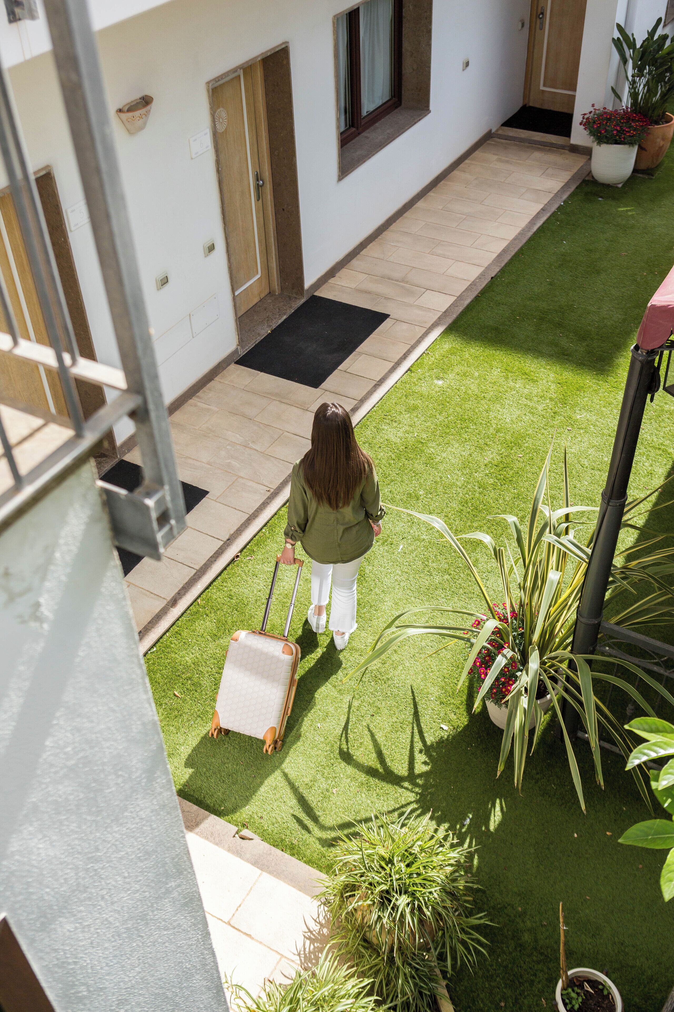 A person walking on a green artificial lawn with potted plants and a suitcase.