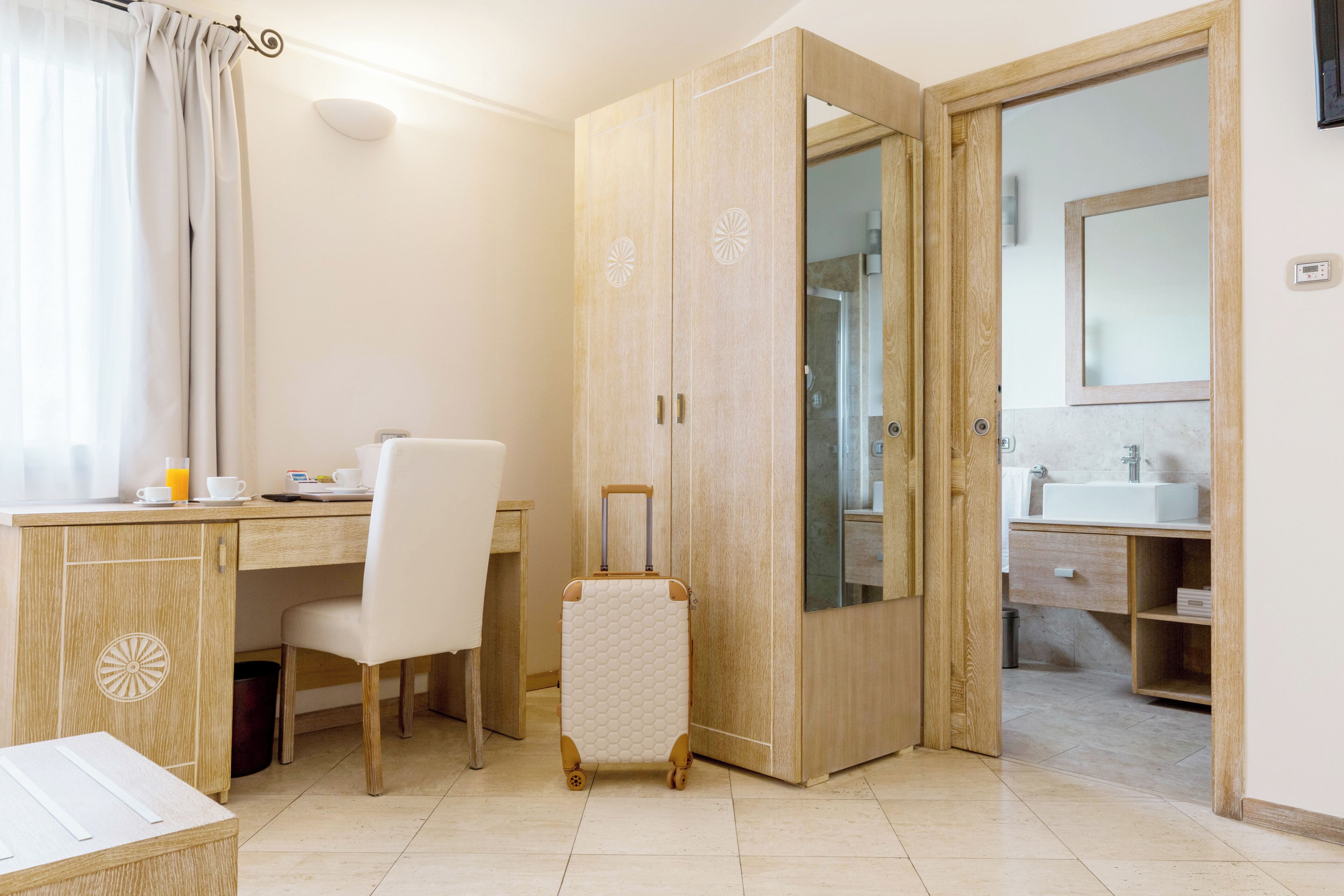 A hotel room with a wooden desk, a chair, a suitcase, and a bathroom with a sink and mirror.